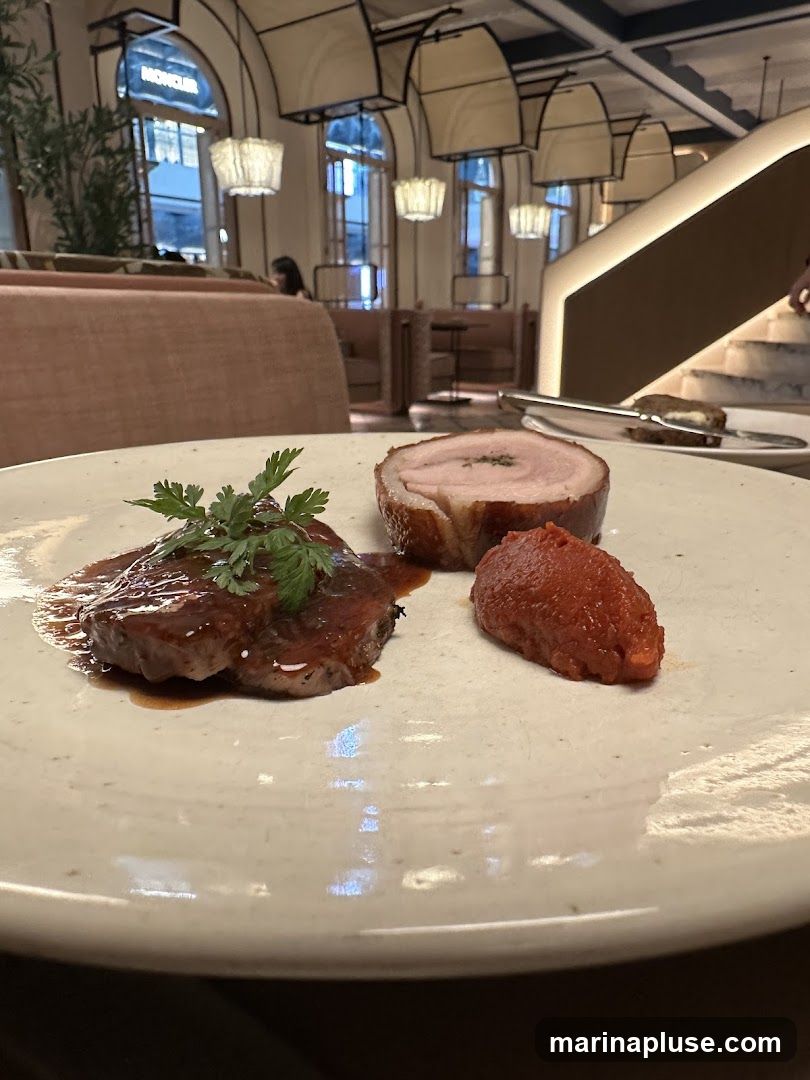 A close-up of a plate with a slice of meat in a dark sauce, a rolled piece of meat, and a dollop of red puree. The background shows a modern restaurant interior with plush seating, decorative lighting, and a staircase. A sign for 'MONCLER' is visible in the distance through a window.