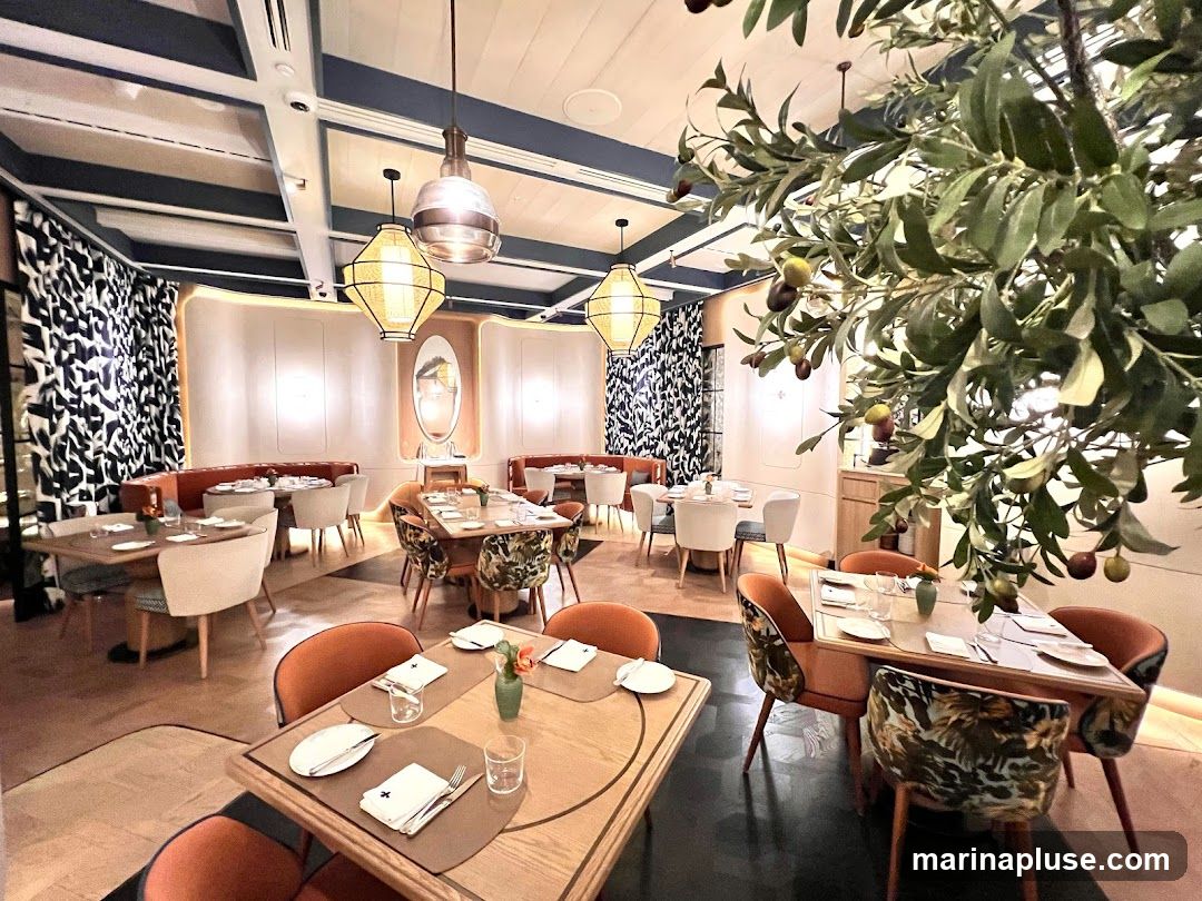 The interior of a modern restaurant with elegant decor. Tables are set with white linens and place settings. Orange upholstered chairs surround some tables, while others have chairs with a tropical leaf pattern. Decorative lighting fixtures hang from the ceiling, and a large artificial olive tree with fruit is a prominent feature. The walls are adorned with patterned curtains and subtle lighting.