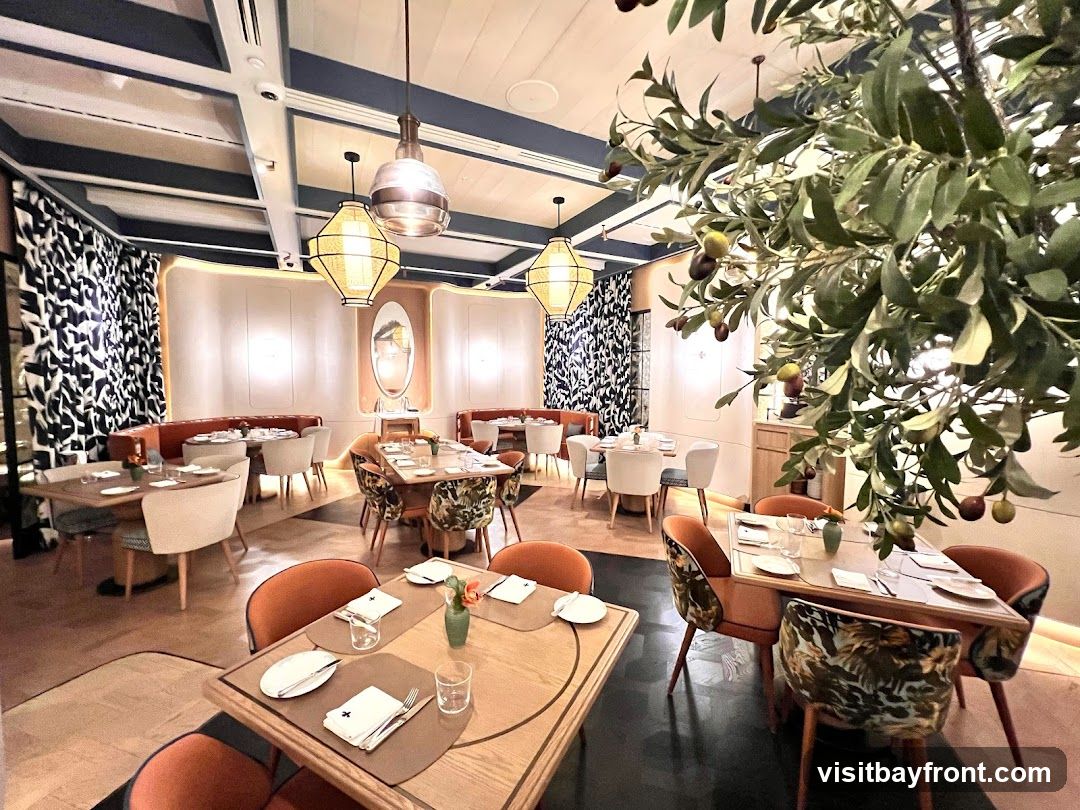 The interior of a modern restaurant with elegant decor. Tables are set with white linens and place settings. Orange upholstered chairs surround some tables, while others have chairs with a tropical leaf pattern. Decorative lighting fixtures hang from the ceiling, and a large artificial olive tree with fruit is a prominent feature. The walls are adorned with patterned curtains and subtle lighting.