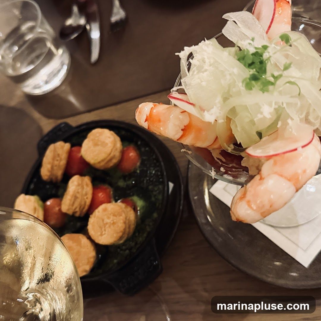 A close-up view of a table setting with two dishes. In the foreground, a glass of white wine is partially visible. To the left, a black cast-iron dish contains small, round pastries or crackers arranged around cherry tomatoes in a green sauce. To the right, a glass bowl holds a shrimp cocktail, featuring several large shrimp served with shredded lettuce and radish slices. The table appears to be made of wood.