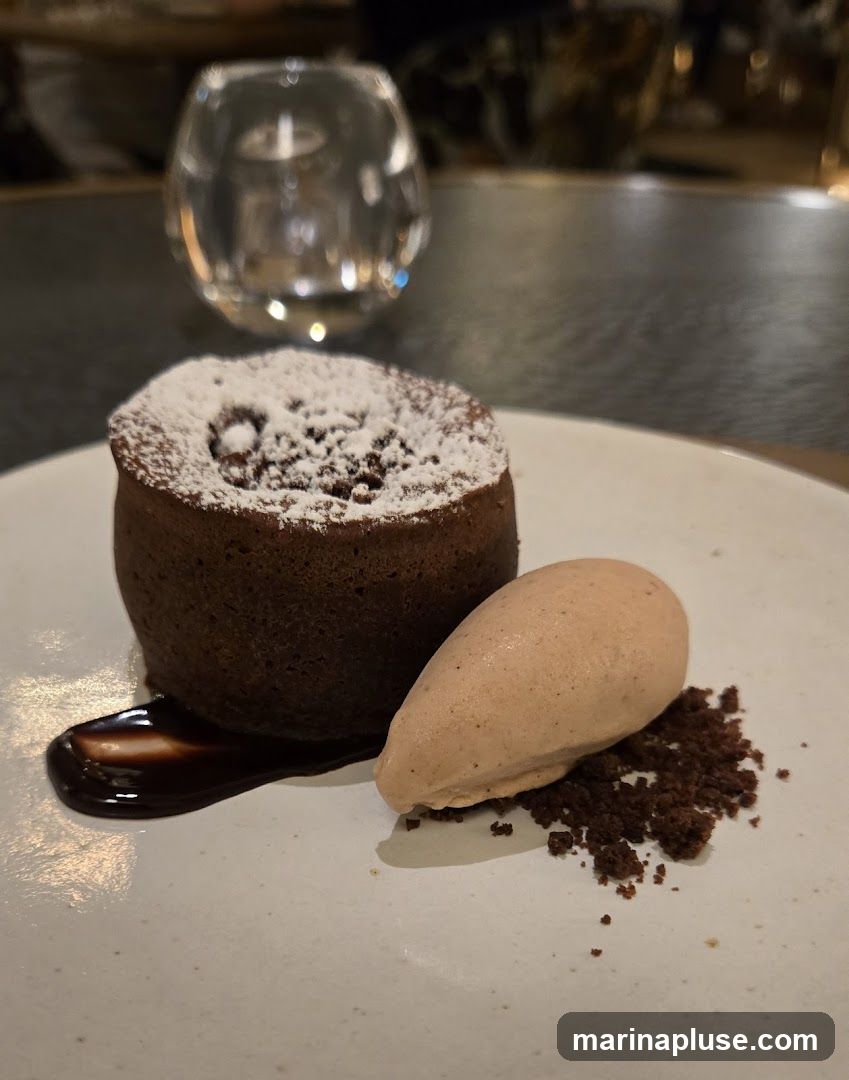 A close-up shot of a chocolate lava cake dusted with powdered sugar, served on a white plate with a scoop of light brown ice cream and chocolate crumbs. A drizzle of chocolate sauce is on the side. In the blurred background, a glass and ambient lighting are visible.