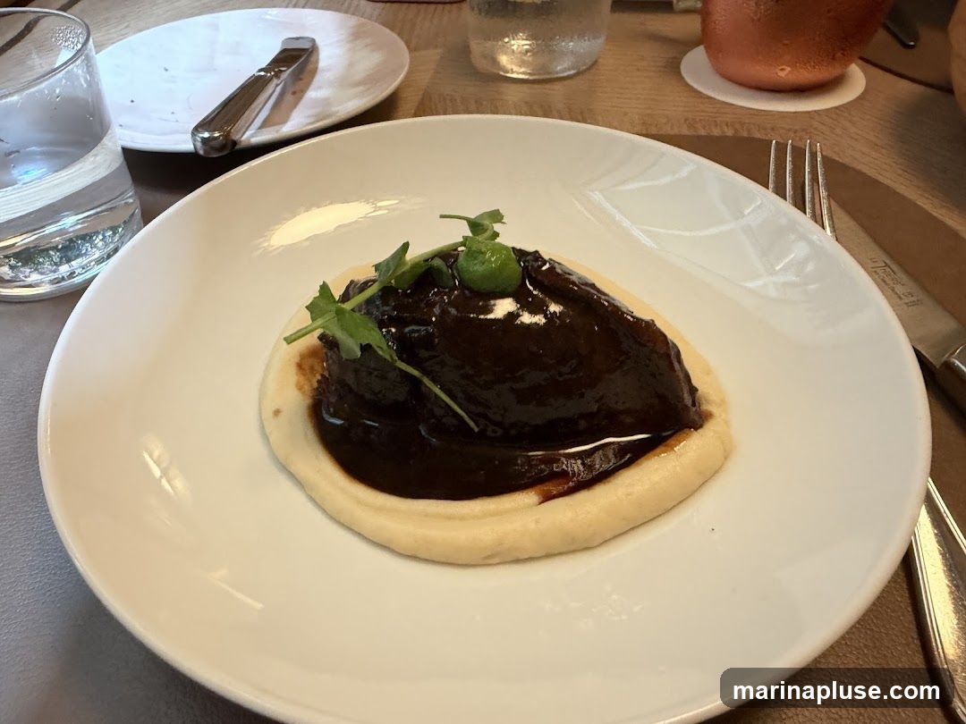 A close-up shot of a white plate holding a serving of braised meat in a dark sauce, presented on a bed of smooth, light-colored puree. The dish is garnished with fresh green herbs and a small green olive or caper. A glass of water and cutlery are visible in the background, suggesting a dining setting.
