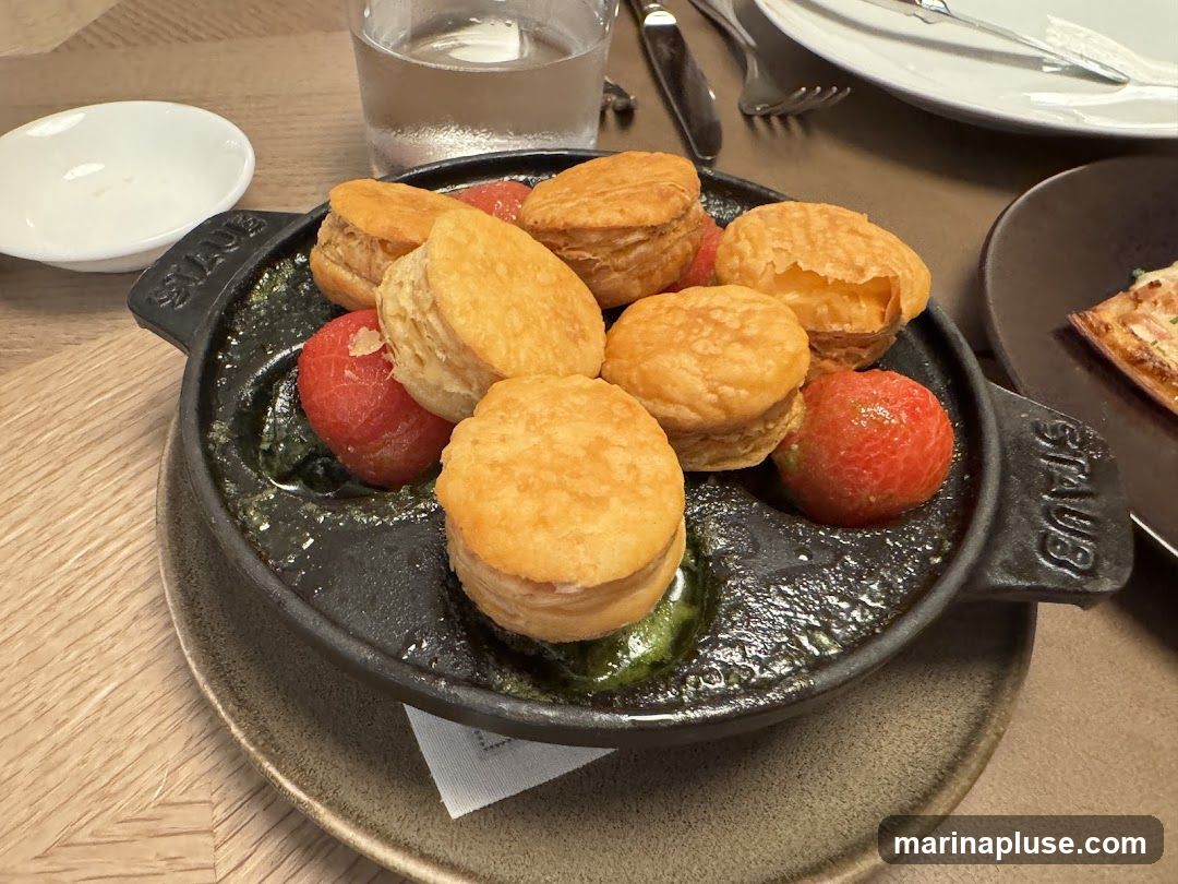 A close-up shot of a dish featuring several golden-brown biscuits served with red berries in a dark cast-iron pan. The pan is filled with a green sauce. A small white dish and a glass of water are visible in the background, along with plates and cutlery.