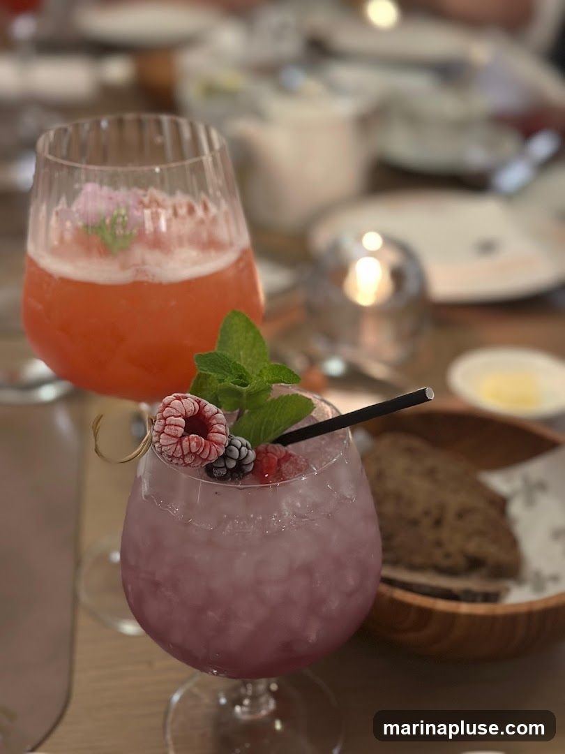 A close-up shot of two colorful cocktails on a table, with a basket of bread in the background. The cocktail in the foreground is a light purple color, garnished with raspberries, a blackberry, and mint leaves. The cocktail in the background is a vibrant pink, topped with foam and a small flower. The table setting suggests a meal or drinks at a restaurant.