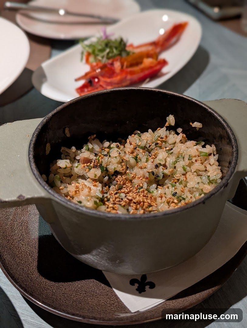 A close-up shot of a bowl of rice topped with sesame seeds and chopped herbs. In the background, a plate holds two cooked shrimp with a small garnish of greens. The setting appears to be a restaurant table.