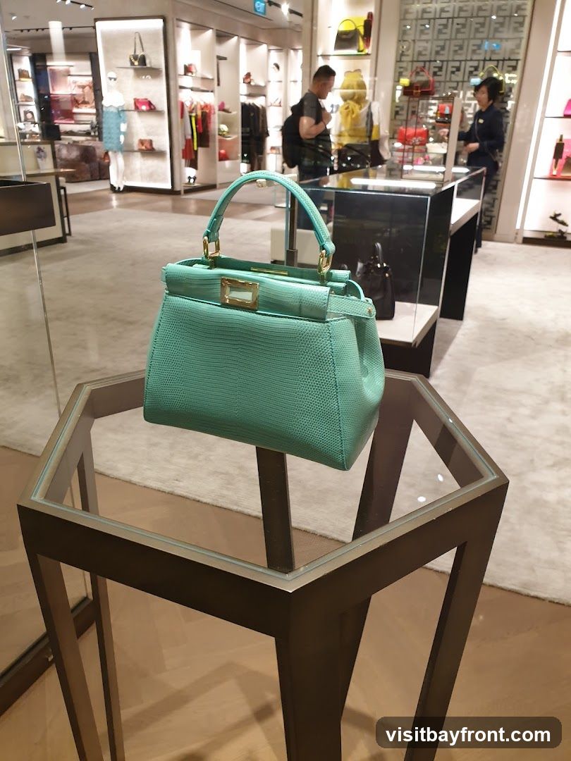 A close-up of a teal-colored handbag with gold hardware, displayed on a hexagonal glass-top table in a luxury retail store. In the background, shelves are stocked with various bags and accessories, and two customers and a sales associate are visible.