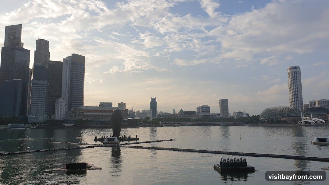 A wide shot of a city skyline across a body of water. The sky is partly cloudy with the sun low on the horizon, casting a warm light. In the foreground, the water has several floating structures, some with lights. The background features a dense collection of modern skyscrapers and other buildings, including a distinctive domed structure. The overall scene suggests an urban waterfront during sunrise or sunset.