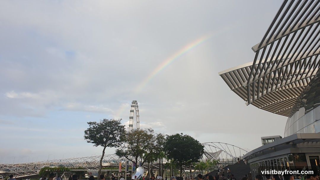 A rainbow arcs across a cloudy sky over a cityscape. In the background, a large Ferris wheel stands tall. In the foreground, a modern building with a distinctive slatted roof is visible, along with a bridge and trees. A crowd of people are gathered in the lower part of the image.