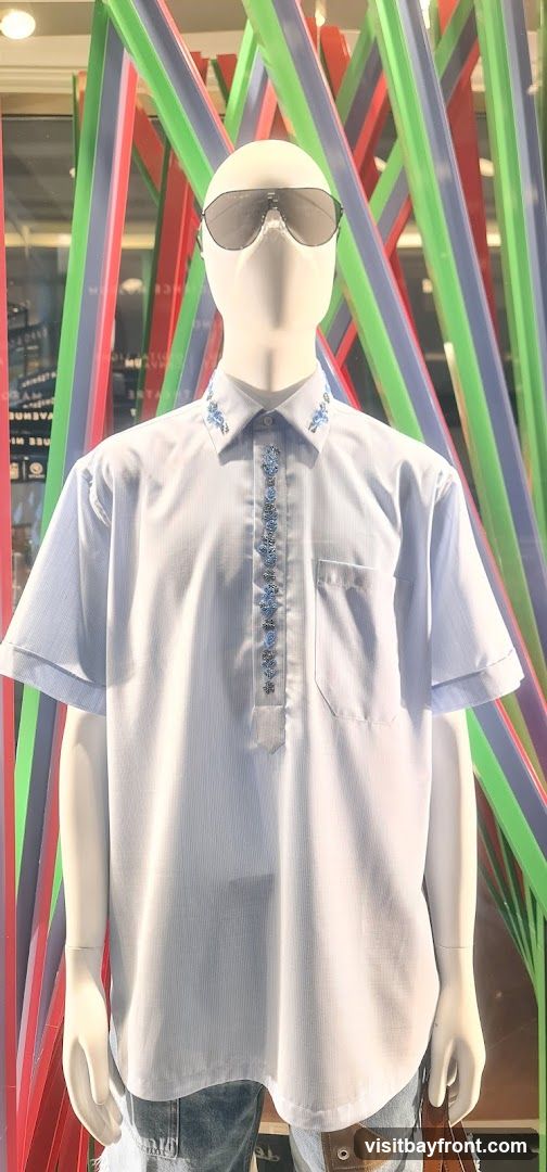 A mannequin is displayed wearing a light blue pinstripe short-sleeved shirt with blue floral embroidery on the collar and down the placket. The mannequin is also wearing sunglasses and denim jeans. Behind the mannequin, colorful vertical stripes of red, blue, green, and purple are visible, suggesting a store display.