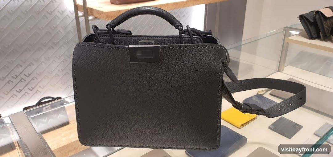 A black pebbled leather handbag with a top handle and a detachable shoulder strap is displayed in a store. The bag has prominent stitching along its edges and a silver-toned clasp closure. In the background, other accessories like wallets and small bags are visible on shelves and in display cases. The wall behind the display has a subtle repeating pattern.
