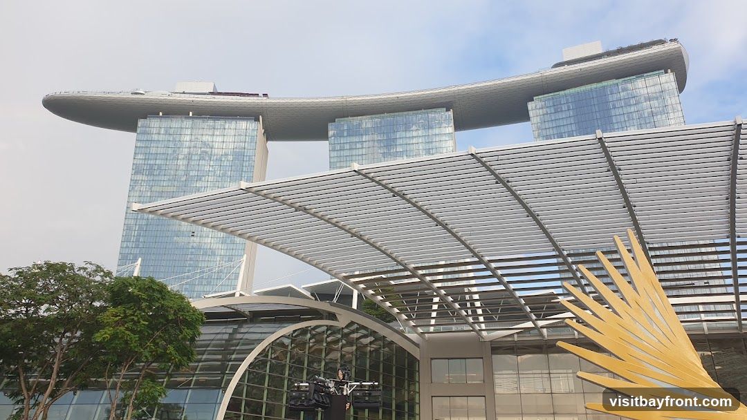A view of the Marina Bay Sands hotel in Singapore, featuring its distinctive boat-shaped top and glass towers. In the foreground, a modern architectural canopy with a golden, fan-like sculpture is visible. Trees and a glass-domed structure are also present.