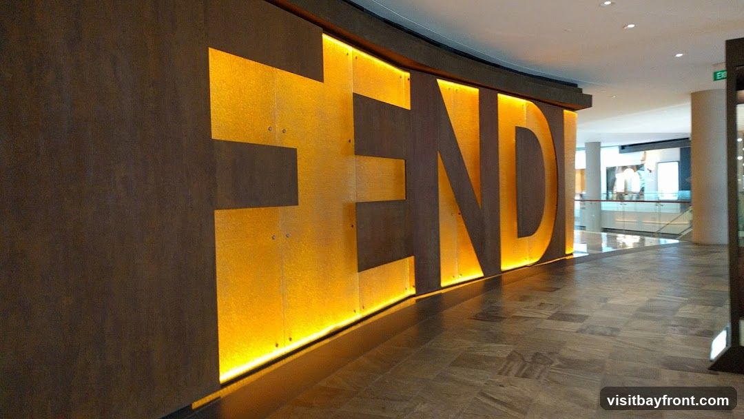 A large, illuminated Fendi logo is displayed on a textured wall inside a modern shopping mall. The logo is made of large, dark letters against a bright, golden-yellow background with warm lighting. The floor is tiled with a grey, stone-like pattern. In the background, other shops and escalators are visible, suggesting a busy retail environment.