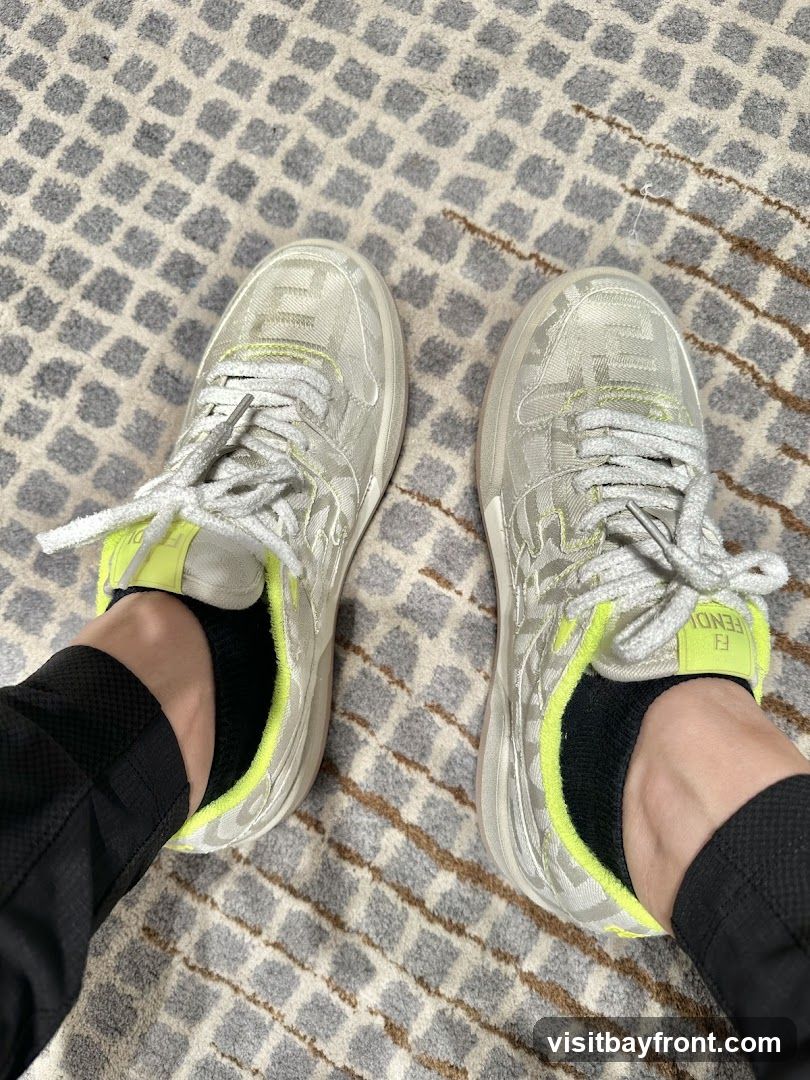 A pair of Fendi sneakers with a white and grey FF motif pattern. The sneakers have white laces and a neon yellow trim around the ankle opening. The Fendi logo is visible on the heel tab. The shoes are worn with black socks and black pants, and are resting on a grey and white patterned rug.