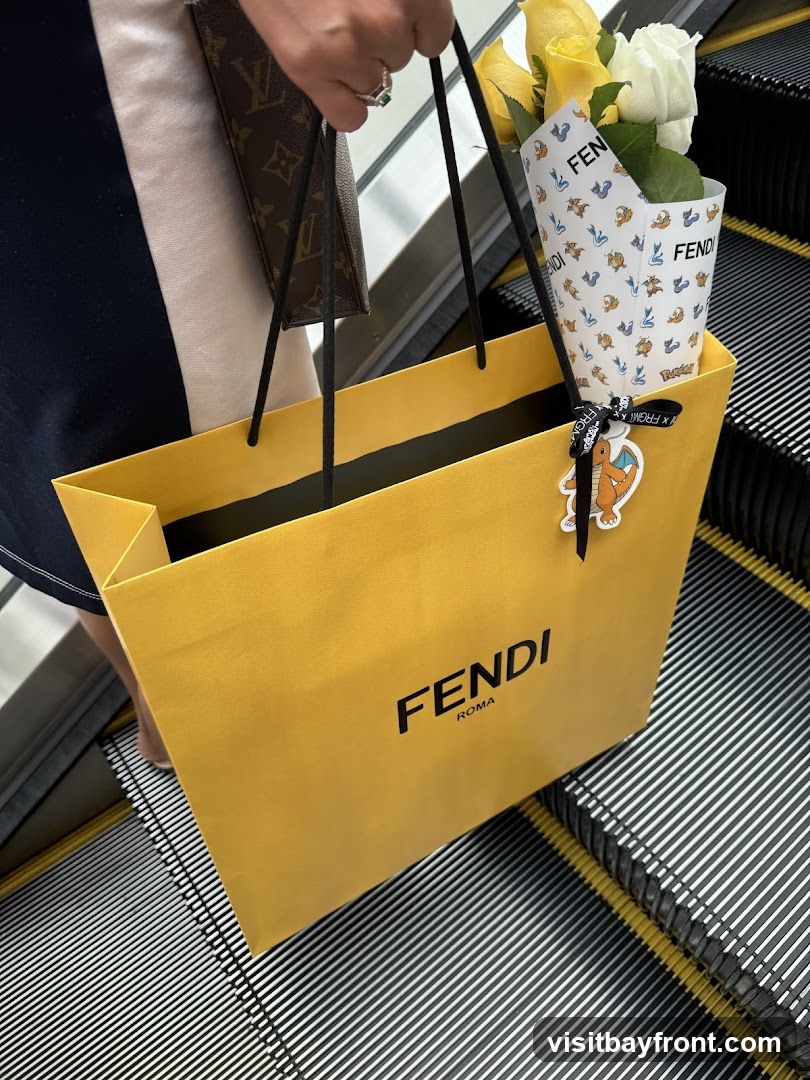 A person is holding a yellow Fendi shopping bag with the Fendi Roma logo on it. The bag is on an escalator. A bouquet of yellow and white roses is tucked into the bag, wrapped in paper with a Fendi logo and cartoon characters. A small charm of a cartoon dragon is attached to the bag's handle.
