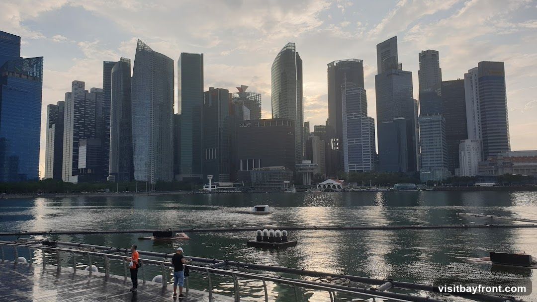 A panoramic view of a modern city skyline with numerous skyscrapers of varying heights and architectural styles, reflected in the calm water below. The sky is partly cloudy with the sun setting or rising, casting a warm glow. In the foreground, a walkway with railings is visible, with two people standing and looking out at the view. Several floating structures are present in the water.