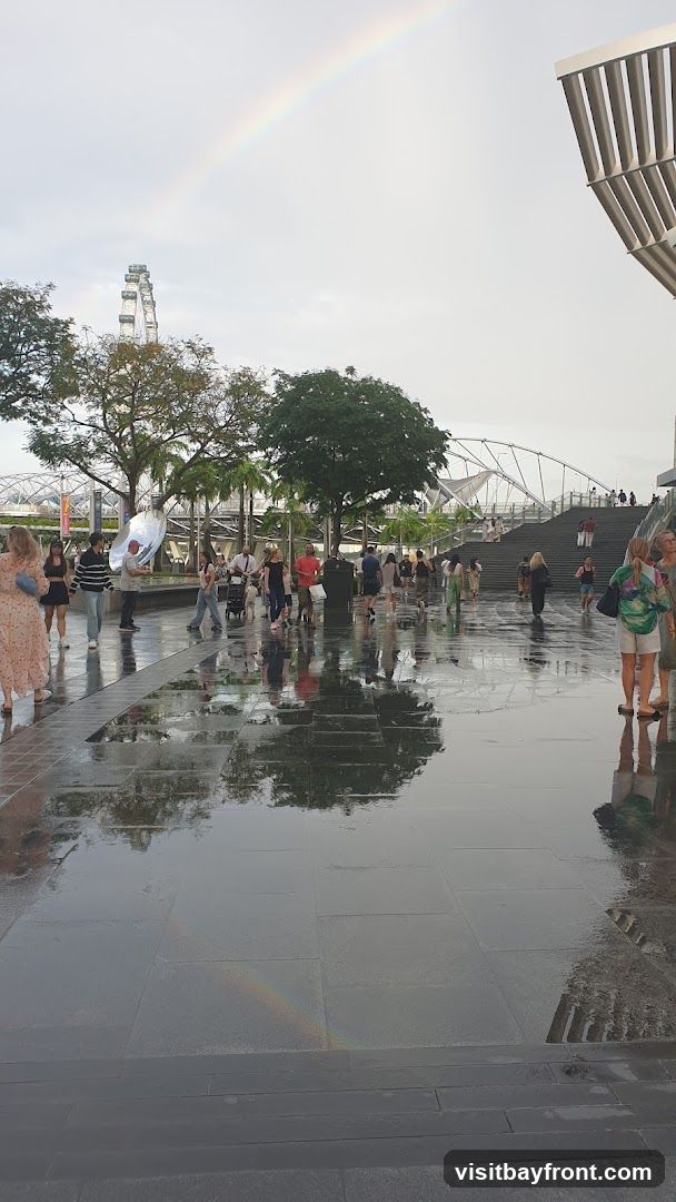 A double rainbow arcs across a cloudy sky over a plaza where people are walking. The ground is wet and reflective, showing the trees and buildings. In the background, a Ferris wheel and a modern bridge are visible.
