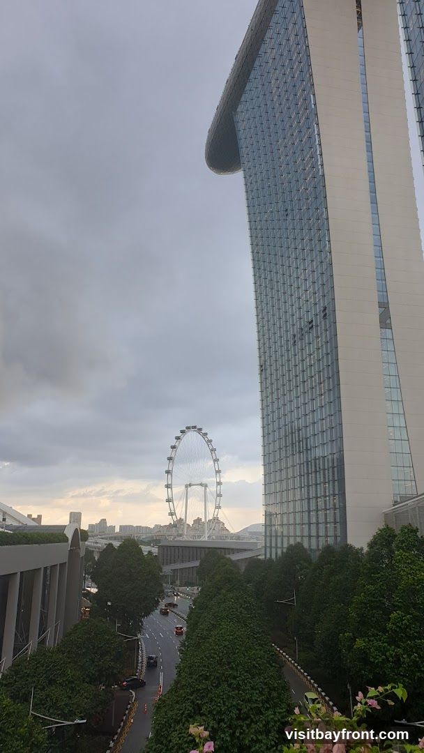 A view of the Marina Bay Sands hotel and the Singapore Flyer observation wheel under a cloudy sky. The hotel's distinctive cantilevered top is visible, along with its glass facade. A road with cars runs through a green, tree-lined area in the foreground.