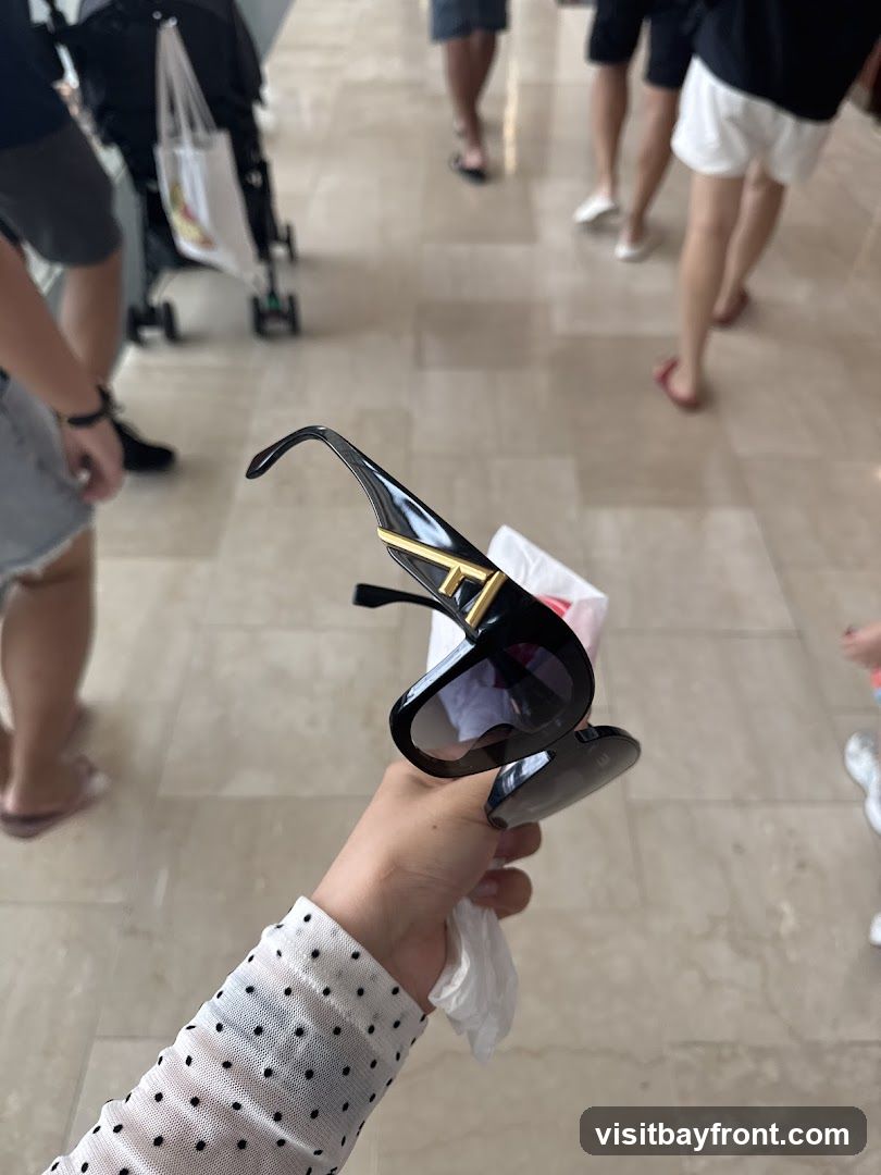 A person's hand holds a pair of black sunglasses with gold accents. The background shows a tiled floor and the legs of several people walking. A stroller is also visible.