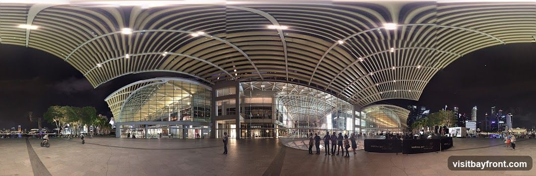 A wide panoramic view of the exterior of Marina Bay Sands at night. The distinctive arched roof structure is illuminated, casting light on the glass-fronted building. People are visible walking and cycling in the plaza area. In the background, the Singapore skyline is visible with its illuminated skyscrapers. Several shops are visible through the glass facade, including one with a prominent 'GUCCI' sign.