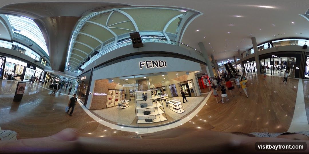 A wide-angle view of a Fendi store located in a modern shopping mall. The store's prominent "FENDI" signage is visible above the entrance. Inside, display shelves showcase handbags and accessories. Other luxury brand stores, such as Dior, are also visible in the background, along with shoppers walking through the mall. The mall features a polished wooden floor and a high, arched ceiling with modern lighting.
