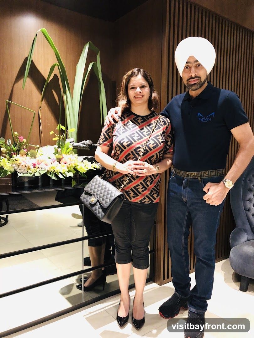 A man and a woman are posing for a photo in what appears to be a high-end interior. The woman is wearing a patterned top and black pants, carrying a quilted handbag. The man, wearing a turban and a dark polo shirt with a logo, is standing next to her. Behind them, there is a decorative arrangement of plants and flowers on a reflective surface, and a wall with vertical wooden panels. A plush armchair is visible to the right.