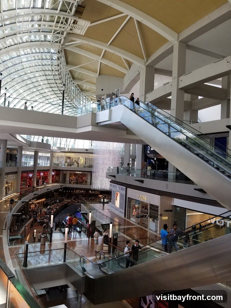 A wide-angle view of the interior of a multi-level shopping mall. The architecture features a large, curved glass ceiling, exposed beams, and multiple escalators connecting different floors. Shops are visible on various levels, including high-end brands like Fendi and Dior. People are seen shopping, walking, and using the escalators. A large, sparkling chandelier-like installation hangs in the central atrium.