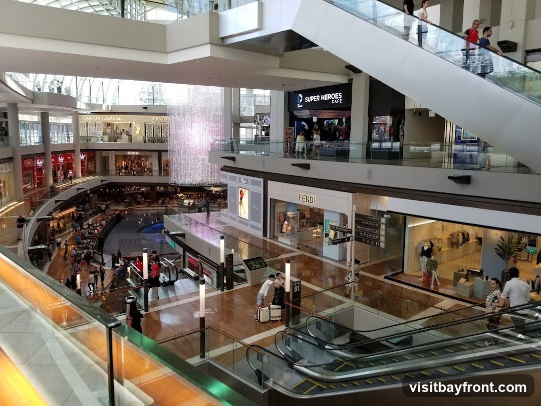 A wide shot of the interior of a multi-level shopping mall. Escalators and walkways connect different floors, with various shops visible on each level. A large, sparkling chandelier hangs from the ceiling in the center of the atrium. People are seen shopping, walking, and riding escalators. Brands like Dior, Fendi, and Super Heroes Cafe are identifiable.