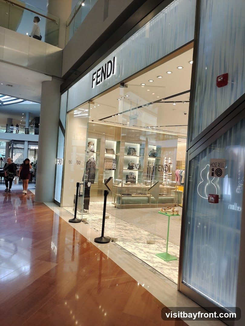 The exterior of a Fendi store in a mall. The Fendi logo is prominently displayed above the glass storefront. Inside, mannequins and displays showcase handbags and clothing. The floor is polished wood, reflecting the store's interior lights.
