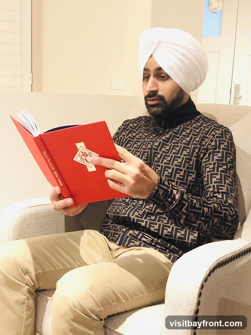 A man wearing a white turban and a patterned jacket sits in a chair and reads a red book. The book has text on the spine that reads "Rocco Rorambini, Bitter Leaves" and a symbol on the cover.