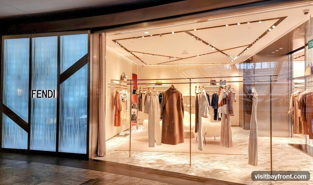 A well-lit, modern storefront with the Fendi logo displayed prominently on a frosted glass panel. Inside, clothing and accessories are neatly displayed on racks and shelves, with a plush seating area visible in the background. The interior features a clean, minimalist design with warm lighting.