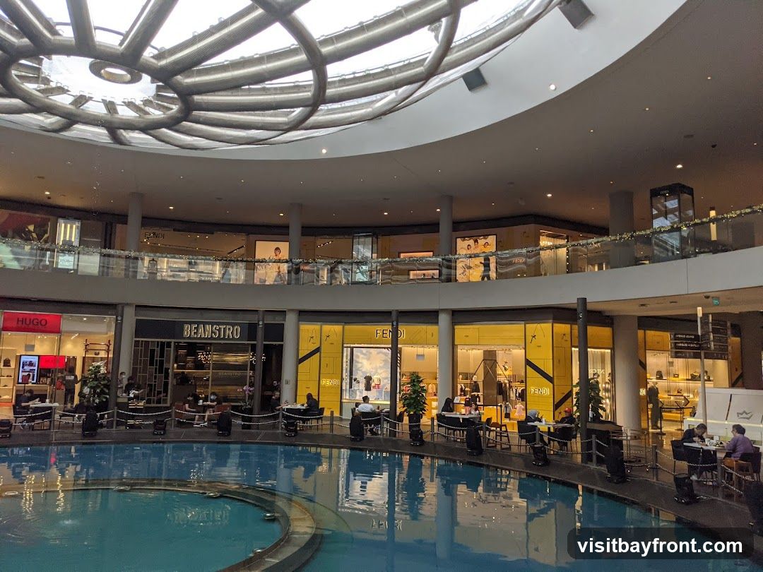 An interior view of a modern shopping mall with a large circular water feature in the foreground. Shops are visible on multiple levels, including Hugo, Beanstro, and Fendi. People are seated at tables around the water feature and inside some of the shops. The ceiling features a distinctive metallic circular design.