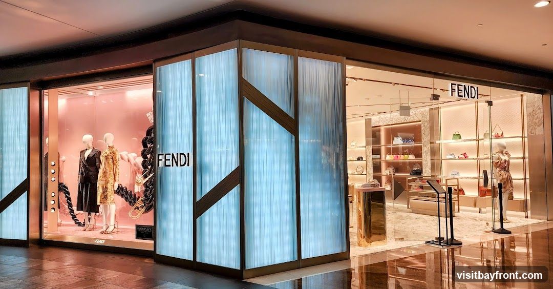 The exterior of a Fendi store with a large display window on the left showcasing mannequins in fashionable attire. The store entrance is on the right, with the Fendi logo prominently displayed above the entrance. The interior of the store is visible, featuring shelves stocked with handbags and other accessories.