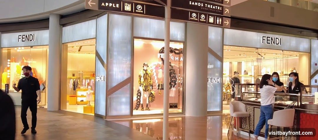 The exterior of a Fendi store with large display windows. Mannequins are dressed in fashionable clothing and accessories. Several staff members are visible inside the store, interacting with customers. Signage for Fendi is prominently displayed above the entrance. Directions to Apple, South, Central, North, and Sands Theatre are also visible.