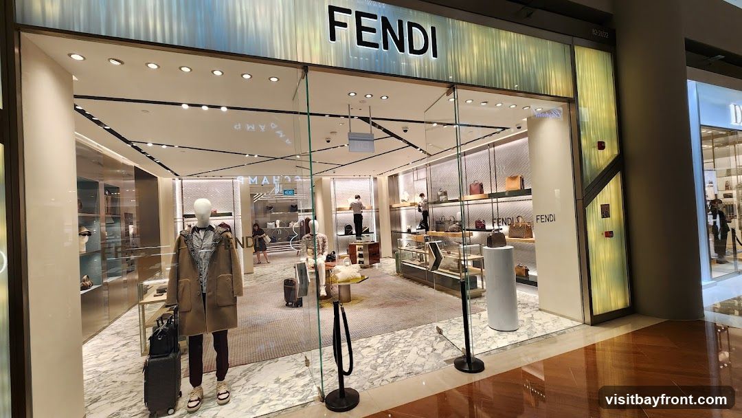 The exterior of a Fendi store with the brand name prominently displayed above the entrance. The store features large glass windows showcasing mannequins dressed in clothing and displays of handbags and accessories. Inside, several staff members are visible attending to the merchandise.
