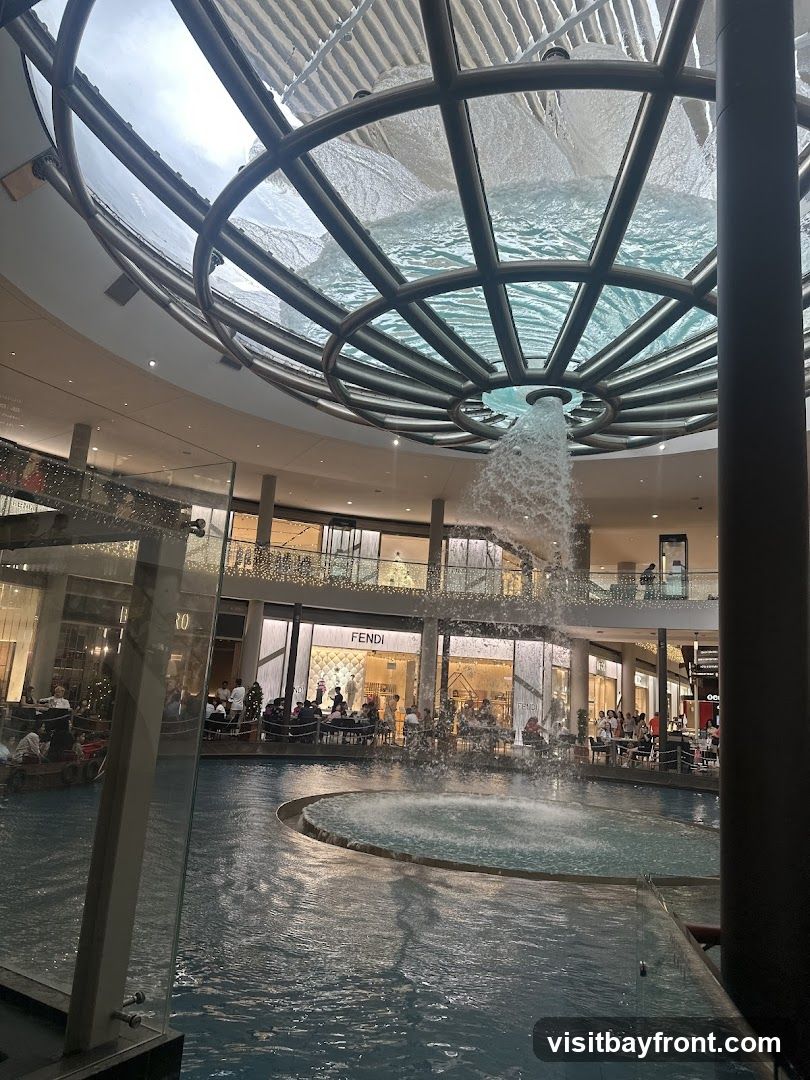 An indoor shopping mall features a large, multi-story waterfall cascading into a pool. Shops with illuminated storefronts, including Fendi, are visible in the background, with people seated at tables in front of some establishments. The mall has a modern architectural design with a glass and metal ceiling structure.