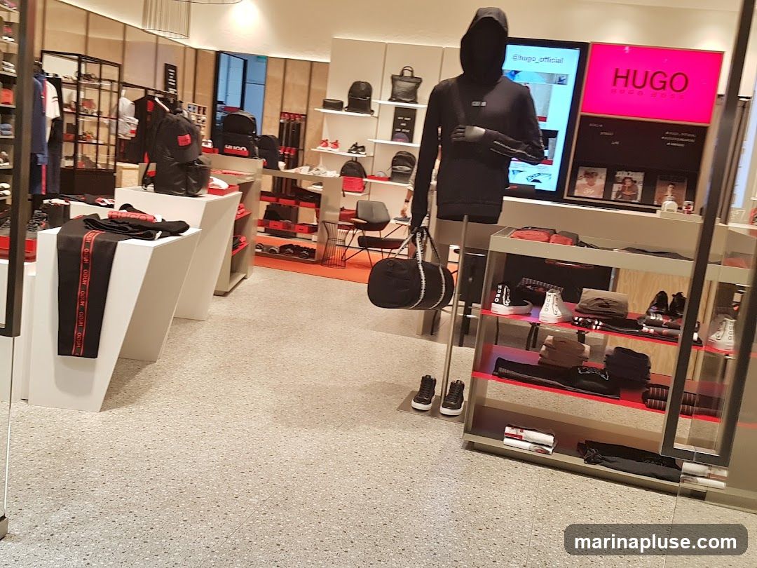 The interior of a Hugo Boss store displays clothing and accessories. Racks of apparel, including pants with prominent branding, are visible. Mannequins are dressed in hoodies and holding bags. Shelving units showcase shoes, bags, and folded clothing. A large screen displays the Hugo Boss logo and social media handle.
