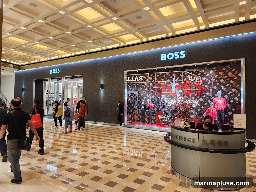 A BOSS store with its prominent logo is visible, featuring a display window with mannequins dressed in red clothing and Looney Tunes characters. To the right, a concierge desk with staff is present. The interior of the mall with its patterned floor and ceiling is also visible.
