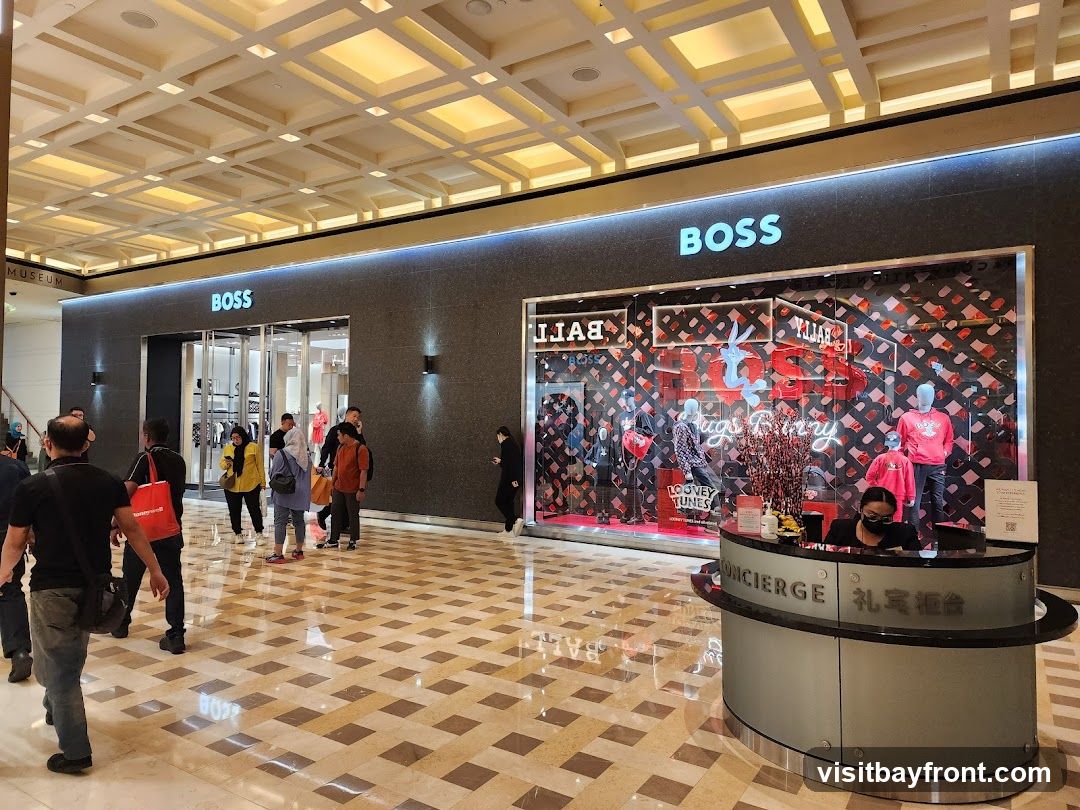A BOSS store with its prominent logo is visible, featuring a display window with mannequins dressed in red clothing and Looney Tunes characters. To the right, a concierge desk with staff is present. The interior of the mall with its patterned floor and ceiling is also visible.