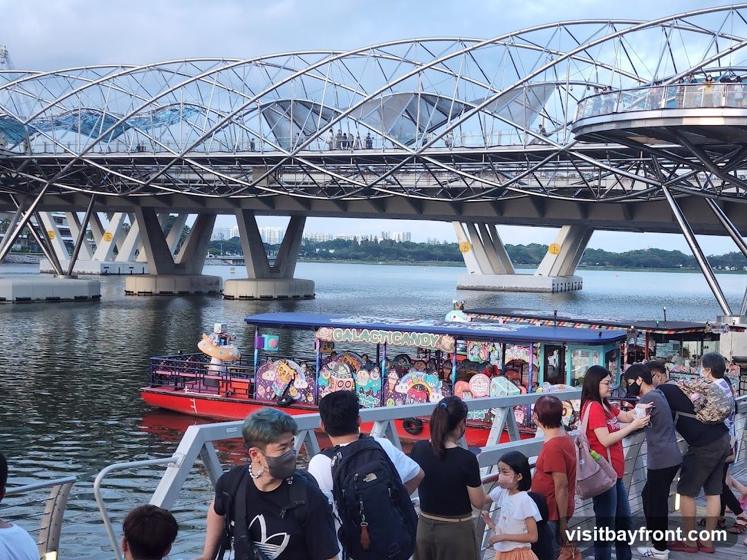 A red boat with a blue canopy is on the water, decorated with colorful cartoon characters and the word "GALACTIC CANDY" in large letters. Several people are standing on a walkway in the foreground, looking towards the boat. A large, modern bridge with a distinctive curved design spans the water in the background. The sky is overcast.