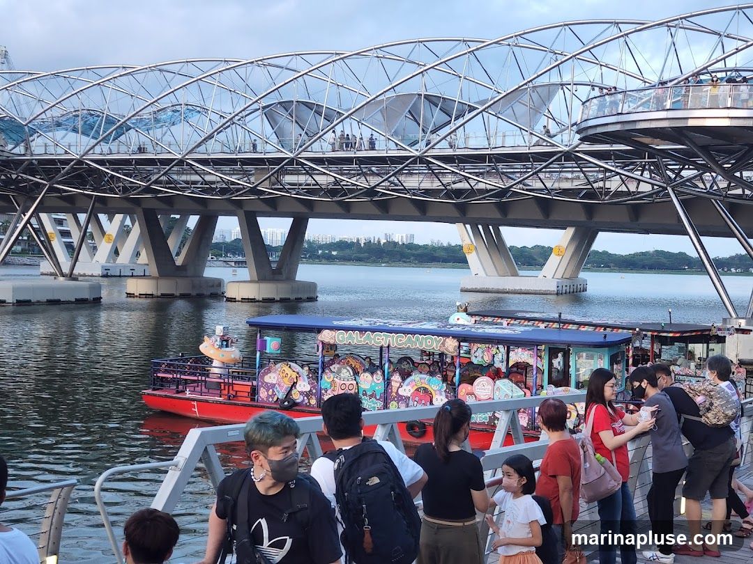 A red boat with a blue canopy is on the water, decorated with colorful cartoon characters and the word "GALACTIC CANDY" in large letters. Several people are standing on a walkway in the foreground, looking towards the boat. A large, modern bridge with a distinctive curved design spans the water in the background. The sky is overcast.