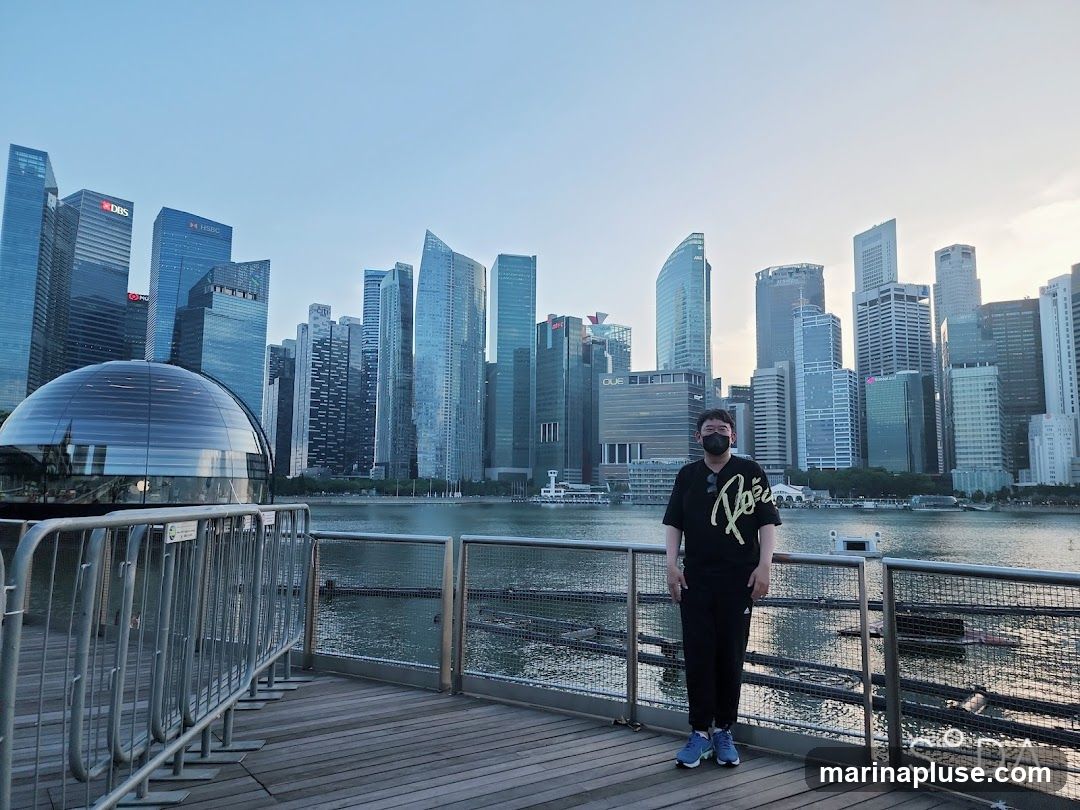 A person wearing a black t-shirt, black pants, and a face mask stands on a wooden boardwalk. Behind them is a body of water and a cityscape with numerous skyscrapers. The sky is clear and blue. A glass dome structure is visible to the left of the person.