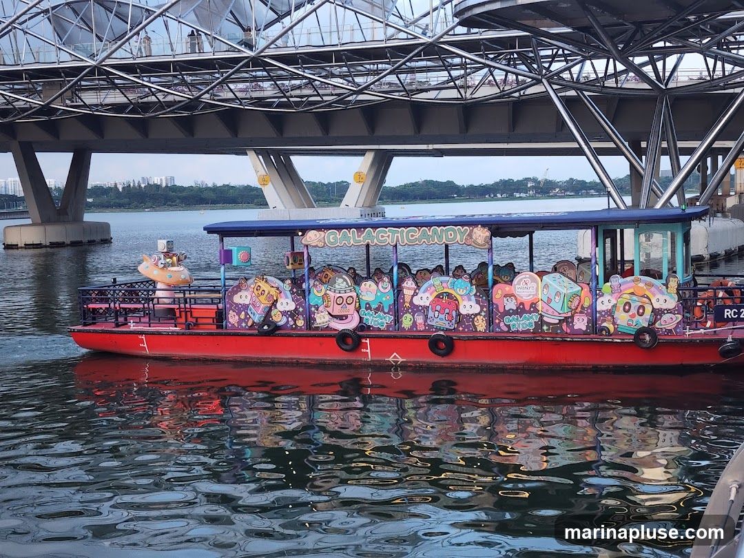 A red boat with a blue canopy is decorated with colorful cartoon characters and the words "GALACTIC CANDY" on its side. The boat is on the water, with a large metal bridge overhead and a cityscape in the background. The boat's decorations appear to be candy-themed, with images of ice cream, lollipops, and smiling characters.