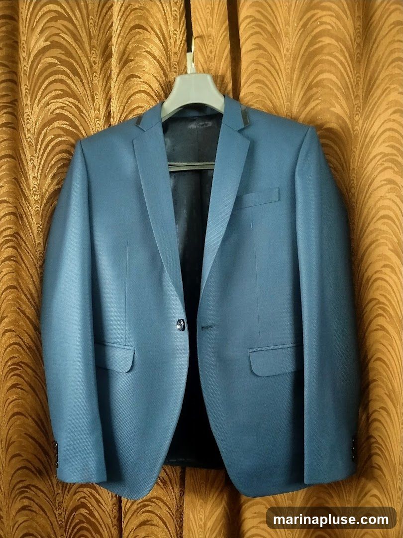A blue blazer is displayed against a backdrop of golden curtains. The blazer has a textured fabric, a single button closure, two flap pockets on the front, and a dark lining.