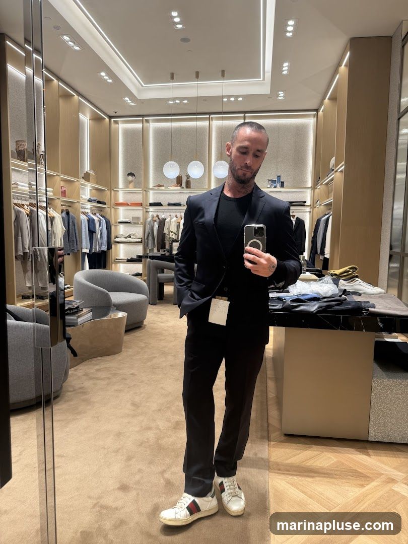 A man in a dark suit, black t-shirt, and white sneakers takes a mirror selfie in a well-lit clothing store. The store features racks of clothing, display shelves with accessories, and modern furniture.