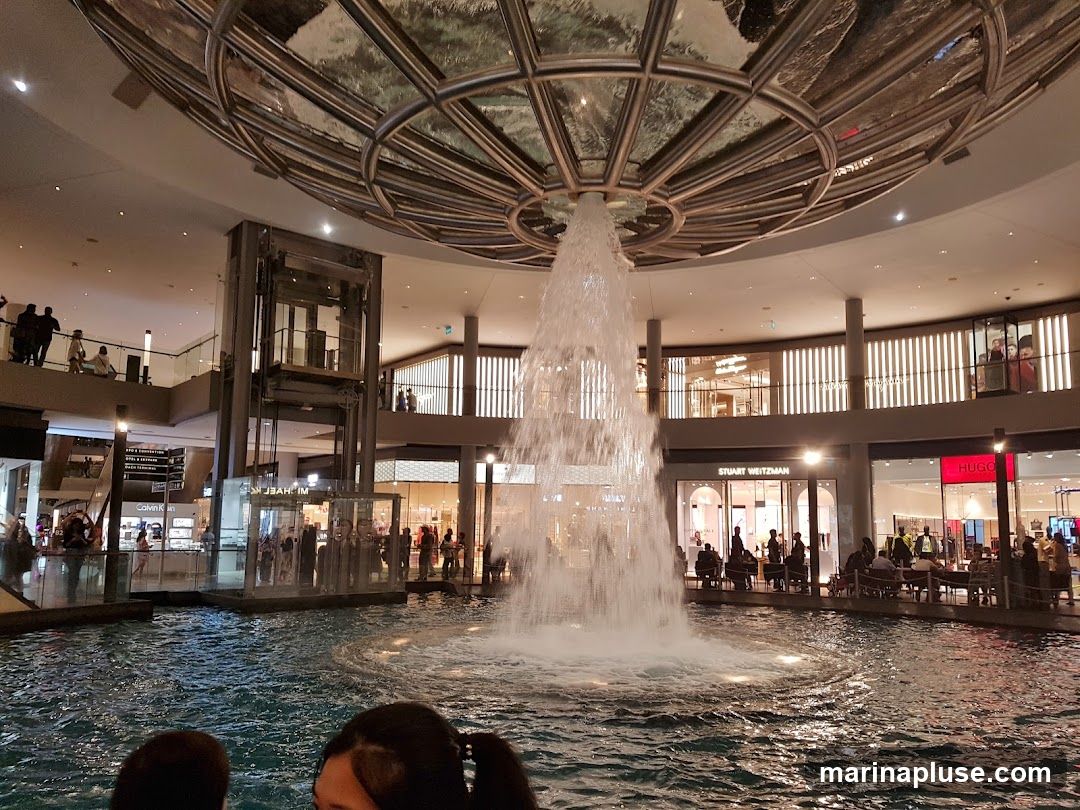 An indoor shopping mall features a large fountain in the center, with water cascading down from a circular structure above. Surrounding the fountain are various shops, including Stuart Weitzman and Hugo Boss, with people browsing and sitting at tables. An elevator is visible in the background, and multiple levels of the mall are populated with shoppers.