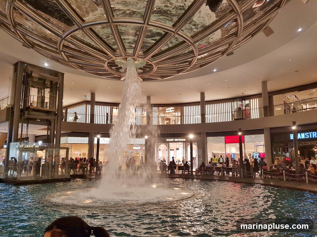 An indoor shopping mall features a large water fountain in the center, with various shops and restaurants visible on multiple levels surrounding it. The architecture includes a domed ceiling with reflective surfaces and escalators. People are seen walking, sitting at tables, and observing the fountain. Visible shop names include Michael Kors, Stuart Weitzman, Hugo, Fendi, and Banstra.