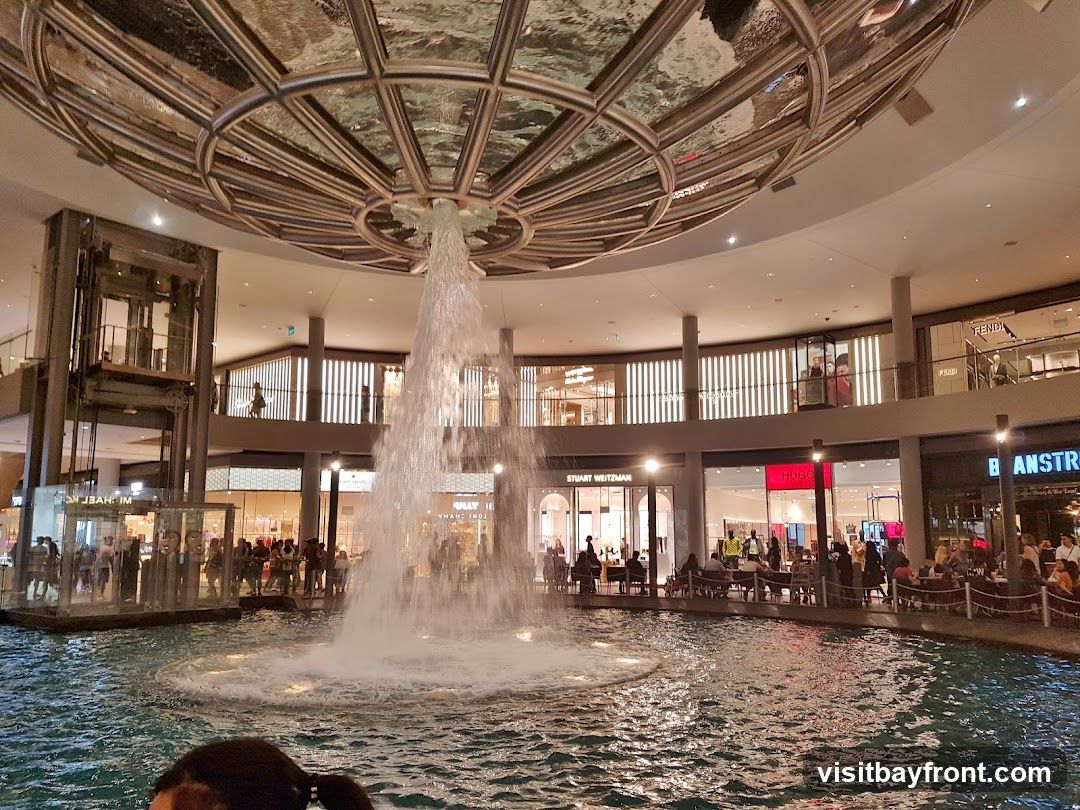 An indoor shopping mall features a large water fountain in the center, with various shops and restaurants visible on multiple levels surrounding it. The architecture includes a domed ceiling with reflective surfaces and escalators. People are seen walking, sitting at tables, and observing the fountain. Visible shop names include Michael Kors, Stuart Weitzman, Hugo, Fendi, and Banstra.