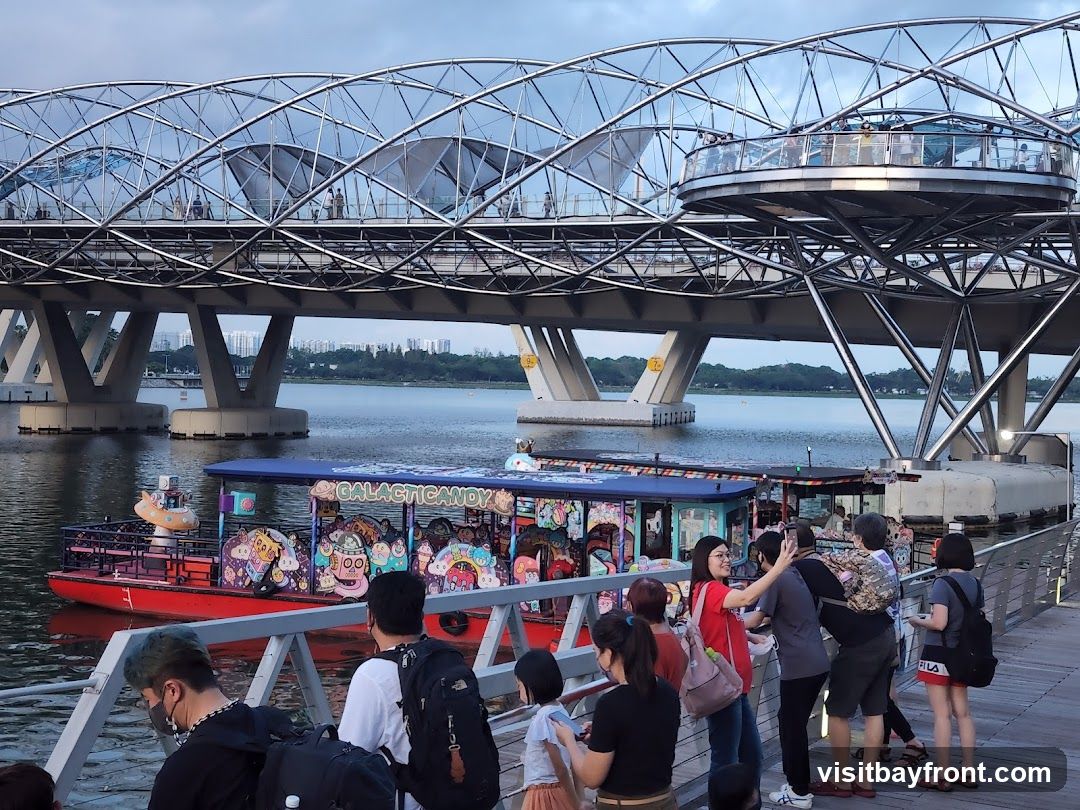 A group of people are standing on a wooden boardwalk, looking at a brightly decorated boat on the water. The boat has cartoonish imagery and the words "GALACTIC CANDY" visible on its side. In the background, a large, modern bridge with a distinctive curved structure spans across the water. The sky is overcast, suggesting it might be late afternoon or early evening.