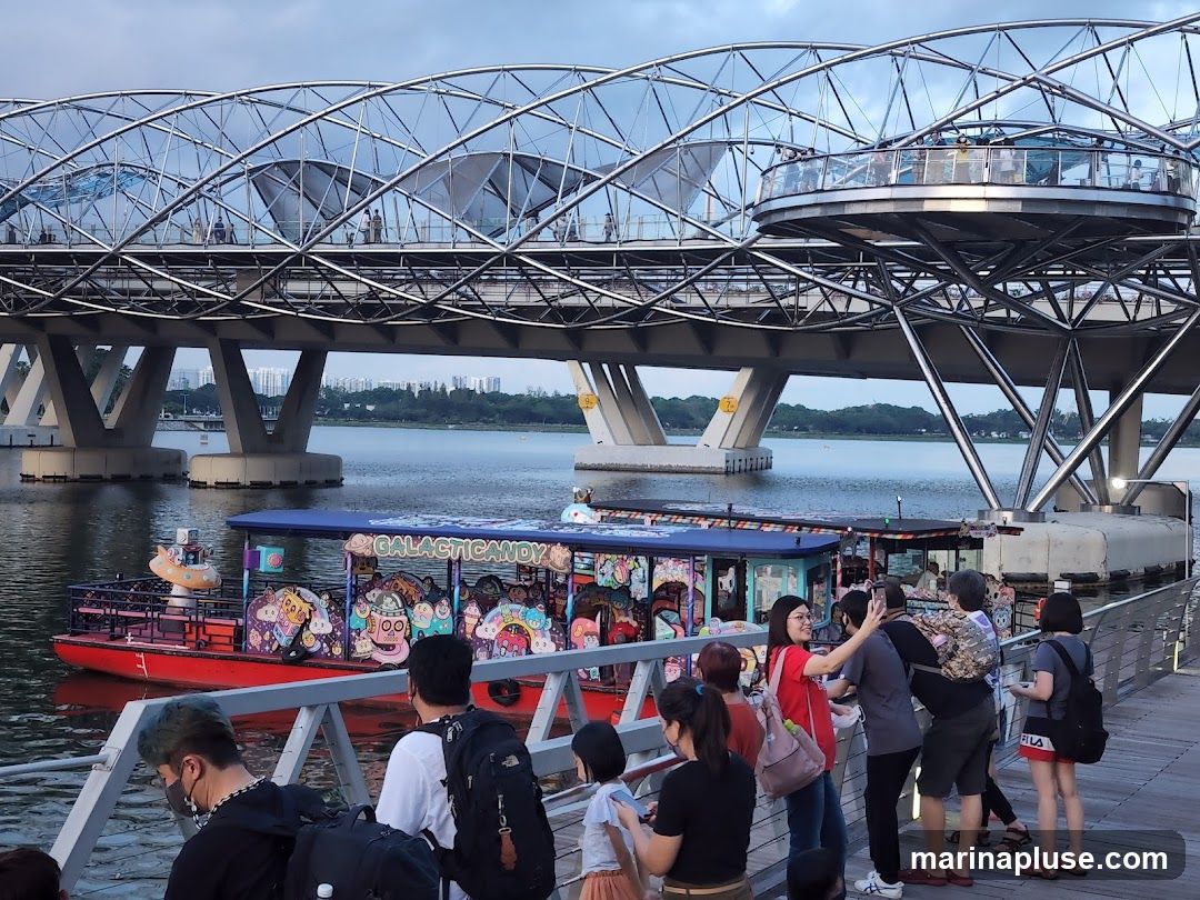 A group of people are standing on a wooden boardwalk, looking at a brightly decorated boat on the water. The boat has cartoonish imagery and the words "GALACTIC CANDY" visible on its side. In the background, a large, modern bridge with a distinctive curved structure spans across the water. The sky is overcast, suggesting it might be late afternoon or early evening.