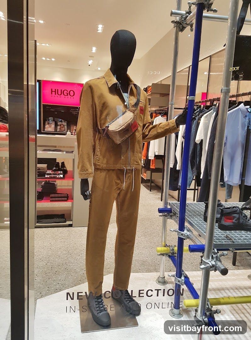 A mannequin dressed in a tan jacket and pants, wearing a crossbody bag, stands in a store window. Behind the mannequin, racks of clothing are visible, along with a large pink sign that reads "HUGO HUGO BOSS". Text on the floor reads "NEW COLLECTION IN-STORE AND ONLINE".